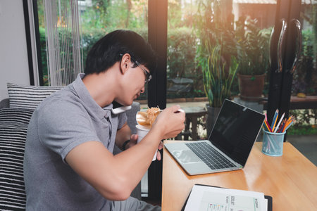 A man who works from home is busy talking on the phone with colleagues about work and eating instant noodles. While his eyes stared at the work content on the laptop as well.の写真素材