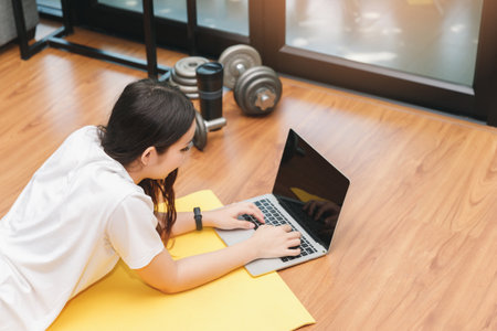 Beautiful young woman watching fitness video tutorials online on laptop while lie down on floor in living room.の写真素材