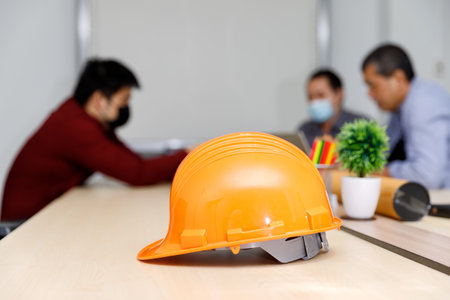 Close up image of a helmet on the desk with architects wearing protective masks working together in the backward, an architectural concept.の写真素材