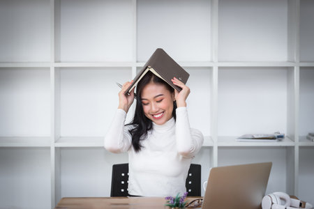 Happy woman sitting at the desk holding a book over her head.の写真素材