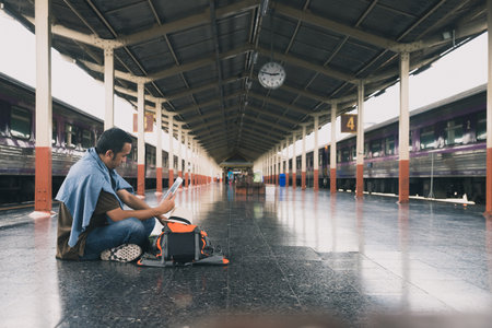 A young man traveler with digital tablet sitting on floor at train station choose where to travel. Concept of backpackers travel adventure by train.の写真素材