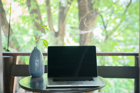 Laptop with a blank screen on a wooden table at home balcony on the background of branches and leaves. Freelance, working from home, online learning, home office. Slow living.の写真素材