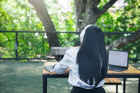 Rear view of Asian woman wearing headphones listening to music on her laptop. Relaxing time. Lazy worker wasting time at workplace in co-working.の写真素材