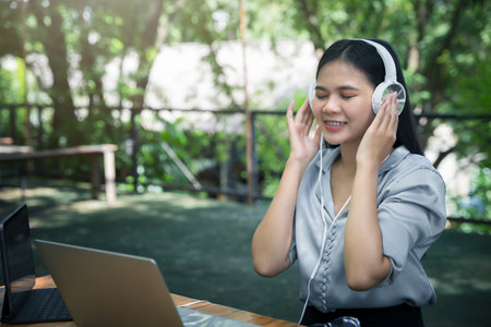 Asian woman wearing headphones listening to music on her laptop. Relaxing time. Lazy worker wasting time at workplace in co-working.の写真素材