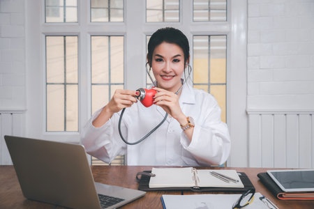 Professional female medical doctor holding a stethoscope check up on a red heart ball in office in...