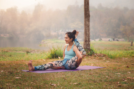 Smiling Asian woman sit down on mat to practicing yoga by raise leg across the occipital in outdoor...
