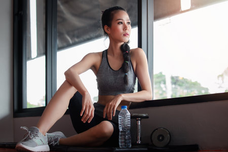 Fitness woman with water bottle and dumbbell sitting on mat by the window and breaking relax after training workout. A woman exercise workout in gym. fitness ,workout, gym exerciseの写真素材
