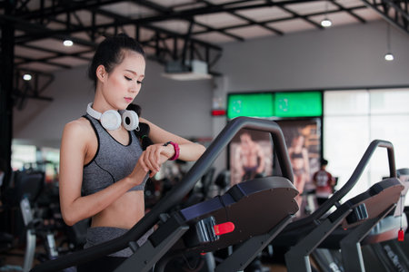Asian woman setting up watch before runing on treadmill at fitness gym club. Sportswoman checking results on smartwatch after running.の写真素材