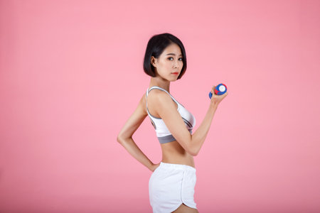 Portrait of a healthy young asian fitness woman doing exercises with dumbbell isolated on pink background. Attractive young asian fitness woman holding dumbell. Sport and healthy lifestyle.の写真素材