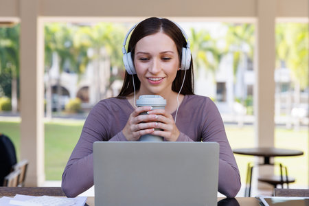 Young female student waering headphones and holding thermos bottle studying with laptop at table in...