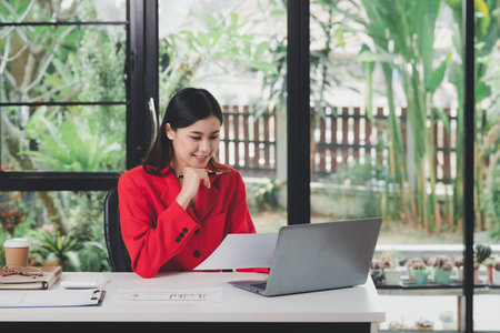 Businesswoman in red suit holding papers preparing report analyzing work results and using laptop. Female executive doing paperwork at workplace using laptop for data analysis.の写真素材