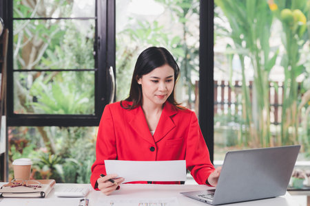 Businesswoman in red suit holding papers preparing report analyzing work results and using laptop. Female executive doing paperwork at workplace using laptop for data analysis.の写真素材