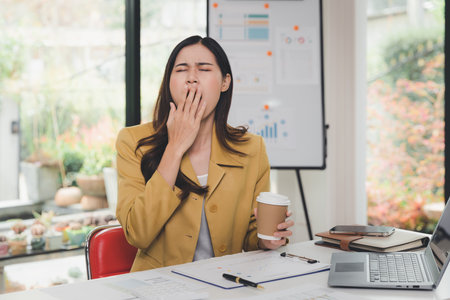 Tired sleepy woman yawning in home office desk and holding a cup of coffee, sleepy, tired, overworked, lazy to work. Overwork and sleep deprivation concept.の写真素材