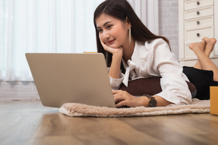 Asia woman lying down on the floor with her legs raised slightly and using laptop at home. Smart working and technology people lifestyle.の写真素材