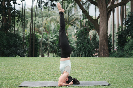 Asian woman doing yoga in Supported Headstand (Salamba Sirsasana) poses on mat in outdoor park.の写真素材