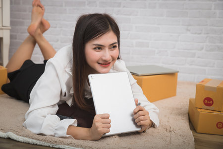 Business woman accepts orders from customer via online sales while lying on floor rug in her home office. Young woman holding tablet happily shopping online.の写真素材