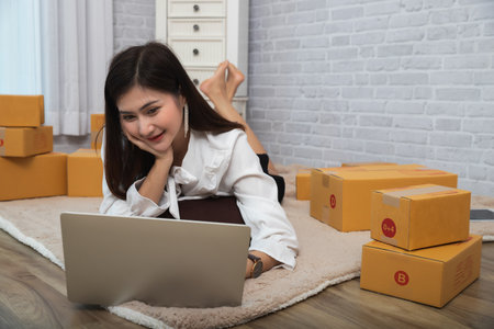 Successful entrepreneur business woman accepts orders from customer via online sales while lying on carpet in her home office. Asian woman working with laptop while lying on carpet at home.の写真素材