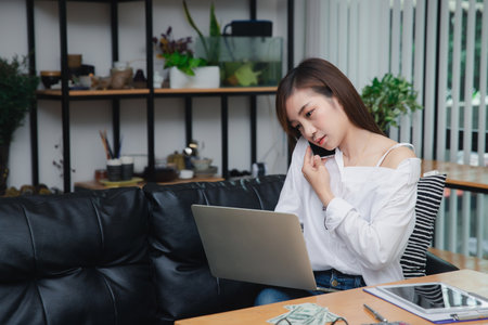 Businesswoman talking on phone working on laptop in modern office. Sharing good business news. Woman working on laptop at office while talking on phone.の写真素材