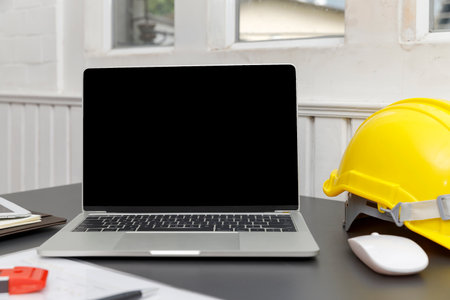 A laptop with a blank screen sits beside a yellow hard hat on a desk, suggesting a blend of technology and construction or engineering.の写真素材