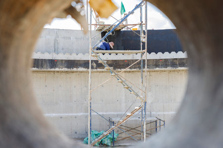 Through the circular frame of a concrete pipe, a construction worker is seen diligently working on scaffolding, representing the meticulous effort behind every structure.の写真素材