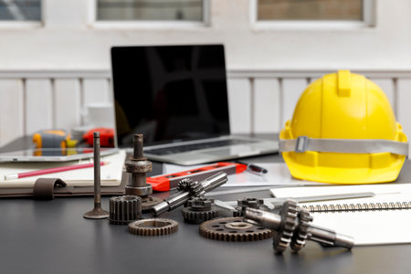 Gear with machine part yellow hard hat sits beside a laptop with a blank screen on engineer desk.の写真素材
