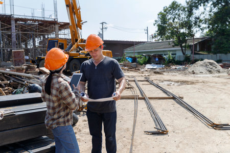 Two construction workers discuss project details on-site, using a tablet and blueprints to plan the next steps amid the bustling construction environment.の写真素材