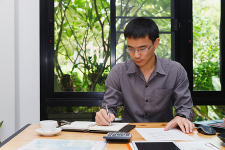 Focused on the task at hand, the individual writes notes with precision, surrounded by documents, a calculator, and a serene environment, making the most of his productive workspace.の写真素材