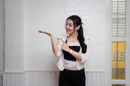 Smiling beautiful Asian woman point to open her empty palm extended sideways presenting copy-space on white wall.の写真素材