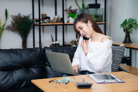 Businesswoman talking on phone working on laptop in modern office. Sharing good business news. Woman working on laptop at office while talking on phone.の写真素材