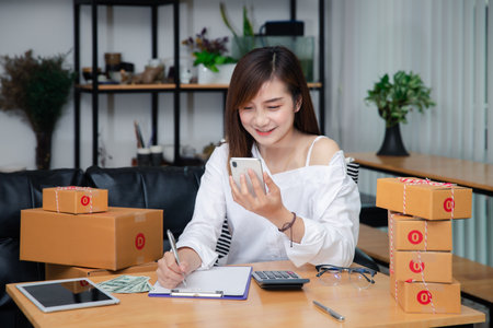 Woman are receiving orders via smartphone. Business woman owner holding parcel box for delivery shipping and using smartphone at home office. Business owner talking on mobile phone and taking order.の写真素材