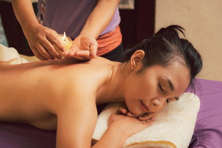 Masseuse drips warm massage oil from a candle on the back of the young woman. Asian woman enjoying back massage with warm massage oil from a candle in massage salon. Beauty treatment concept.の写真素材