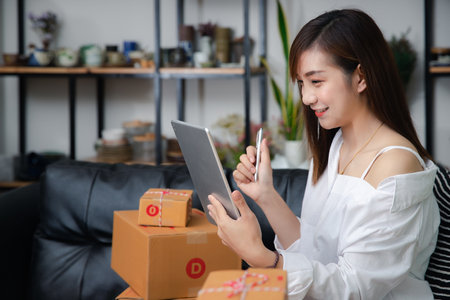 Successful entrepreneur business woman using digital tablet to check and prepare parcel box of product for delivery to customer. Woman with online sales and Parcel shipment in her home office.の写真素材