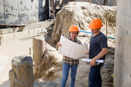 Two construction engineers in hard hats review building plans at an active construction site. An...