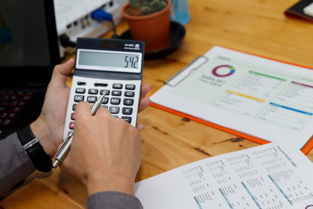 A professional hand calculating numbers with a calculator, pen in hand, while a laptop rest nearby,...