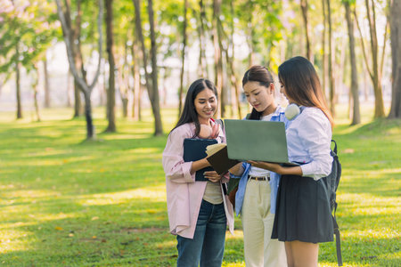 Group of Asian college students standing on green grass and discussing with laptop and books....