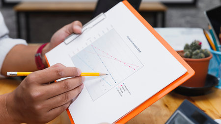 Close-up of hand holding pencil pointing at business analysis chart on clipboard, showing data trend...