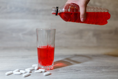 A woman's hand pours a red sports drink or lemonade into a glass Cup from a plastic bottle. Energy drink in glass and plastic bottle on wooden table, scattered pills. Energy for workout in a gym.の写真素材