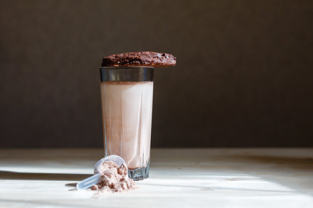 Mixed milk chocolate with whey protein shake in glass cup, scoop with protein and chocolate cookie on wooden table. Dietary snack for bodybuilding. Protein casein, geiner, hydrolysate before trainingの写真素材