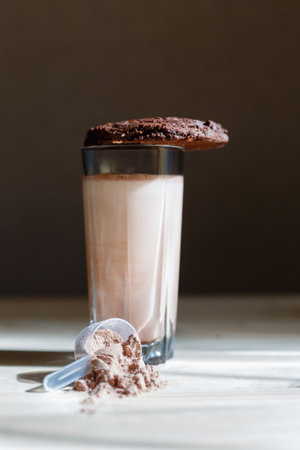 Mixed milk chocolate with whey protein shake in glass cup, scoop with protein and chocolate cookie on wooden table. Dietary snack for bodybuilding. Protein casein, geiner, hydrolysate before trainingの写真素材