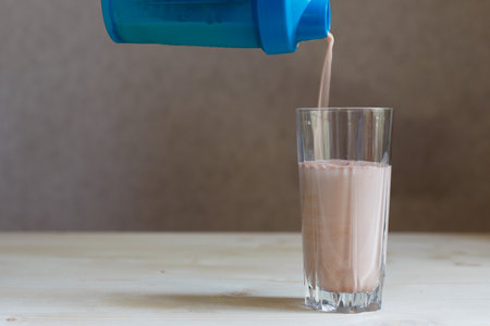 Ready milk whey cocktail protein drink is poured from a shaker into a glass Cup. Sports drink for a set of pure muscle mass. Hand holding plastic shaker with protein shake and glass on tableの写真素材