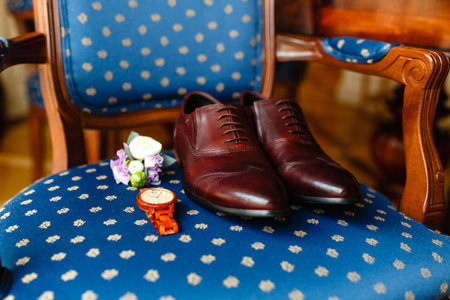 A men's set of accessories on an old wooden chair with a soft seat. The concept of business accessories in clothes. Stylish wristwatch, wooden watch. Men's classic leather shoes, flower boutonniere.の写真素材
