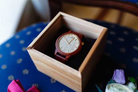 Stylish wristwatch in a wooden box. In background Pink bow tie, beautiful glass cufflinks, flower boutonniere. A men's set of accessories on an old wooden chair with a soft blue seat.の写真素材