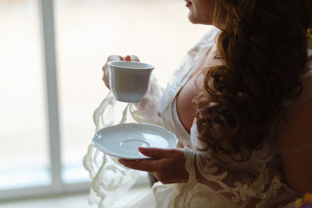 Close-up of a girl's hand in a boudoir dress drinking tea or coffee. The morning of the bride at the hotel before the wedding in the Church.の写真素材