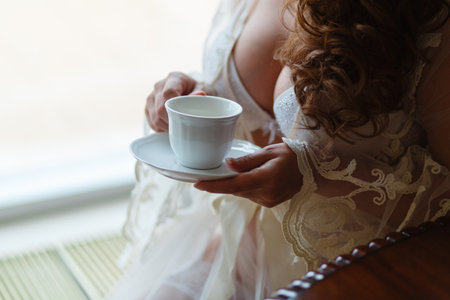 Close-up of a girl's hand in a boudoir dress drinking tea or coffee. The morning of the bride at the hotel before the wedding in the Church.の写真素材