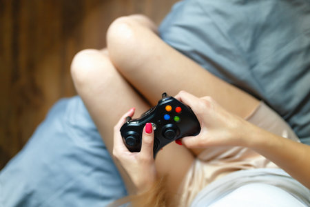Close-up of hand blonde girl with gamepad in hand playing video games console while lying on the bed. The girl is out of focus, holding the joystick of her video console while playing video games.の写真素材