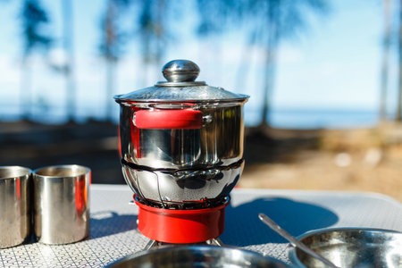 Aluminum pan boil on fire from a portable gas burner next to plates and mugs on a portable table...