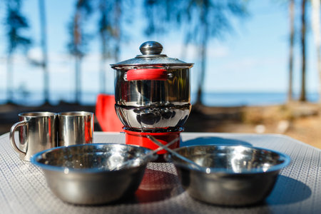 Aluminum pan boil on fire from a portable gas burner next to plates and mugs on a portable table against the background of the red folding chairs in the camping. Cooking while traveling by car.の写真素材