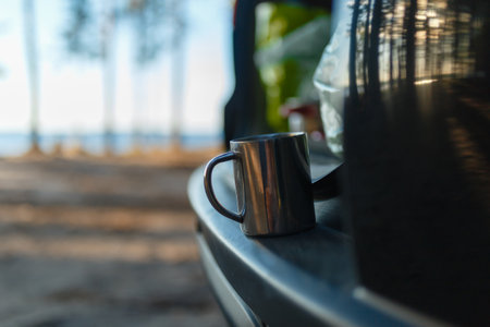 Closeup iron camping mug with a warm drink on the shelf with the open door of the rear trunk of the SUV on the camping background. Travel by SUV or car in the wild. Concept of camping in the forest.の写真素材