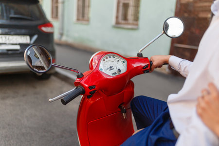 A man driving a red classic scooter rides on the asphalt in the city. Close - up of man's hands holding on to the red scooter's gas handlesの写真素材