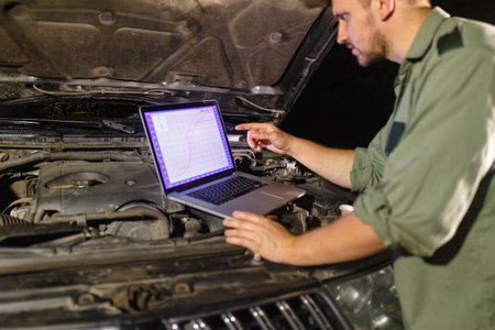 Mechanic in uniform looking intently at the laptop is connected to the motor vehicle for testing and...